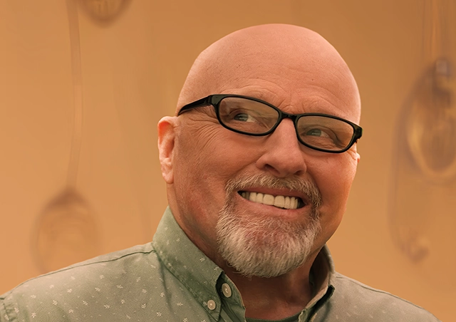 Mickey, an older man with polycythemia vera, smiles in front of a tan-colored, AI-generated backdrop of glass water droplets.